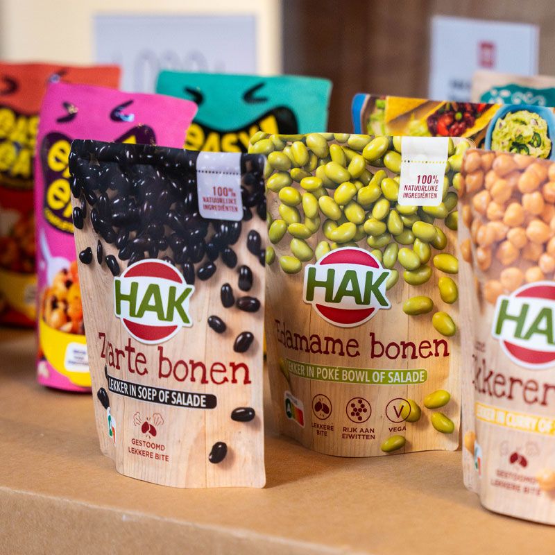 Packs of HAK beans including black, edamame, and chickpeas, displayed on a cardboard surface, showcasing various flavors and uses.