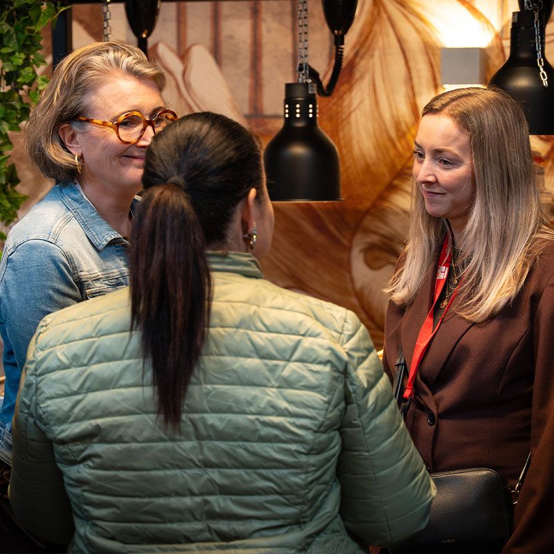Three women engaged in conversation under pendant lights, with one facing away, in a warmly lit room.