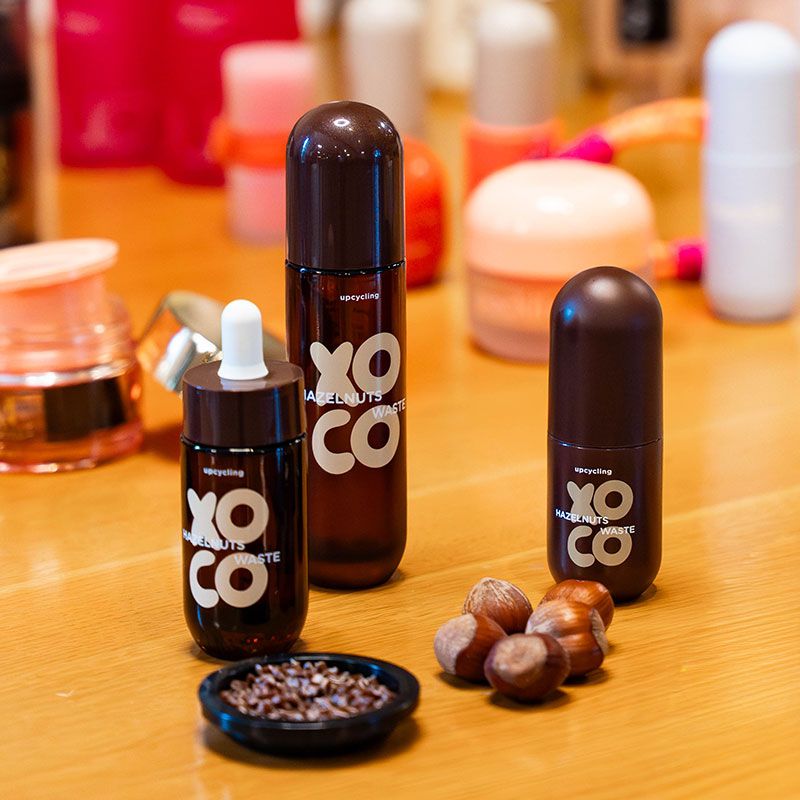 Brown cosmetic bottles labeled "XOCO" on a wooden table with hazelnuts and a small dish of seeds nearby, surrounded by various bottles.