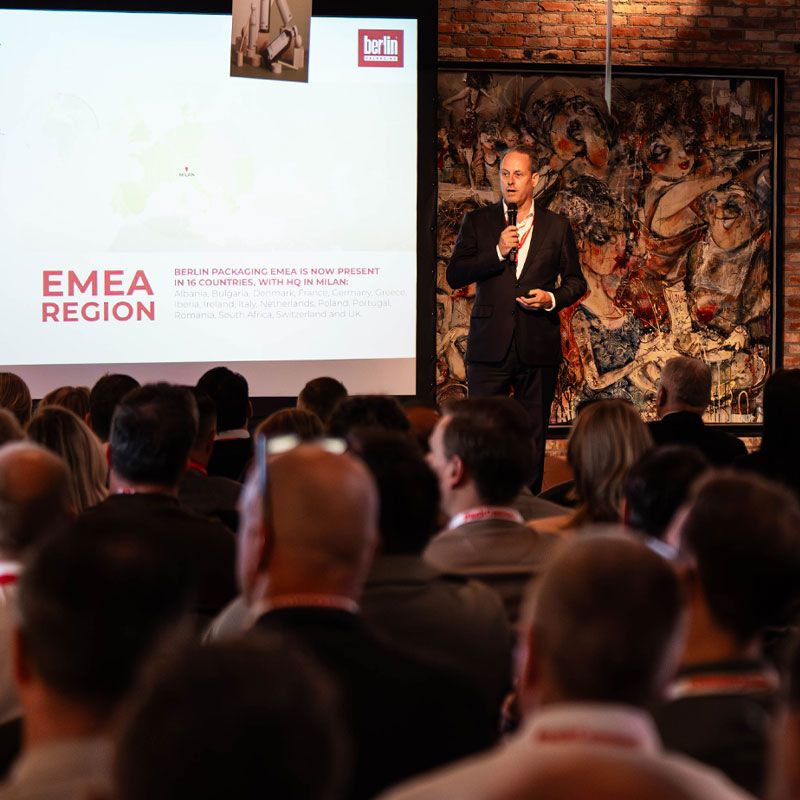 Man in a suit presents at a conference, standing in front of a screen displaying information about Berlin Packaging's EMEA region presence.