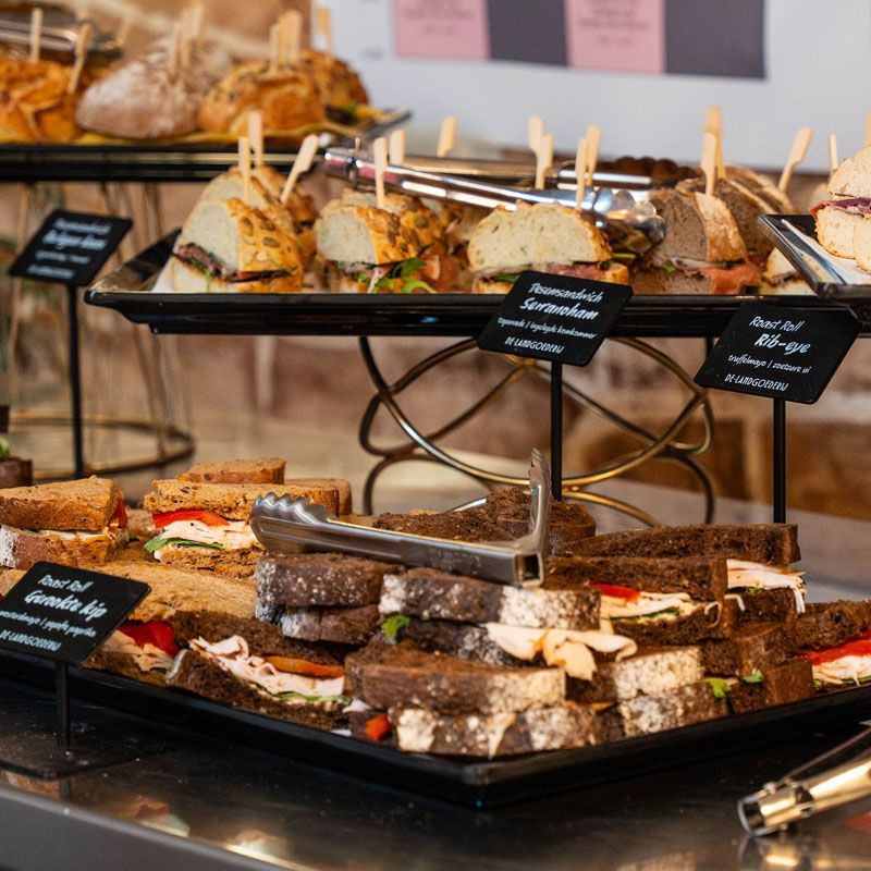 Assorted sandwiches displayed on black trays, with labels and metal tongs, arranged on a buffet table.