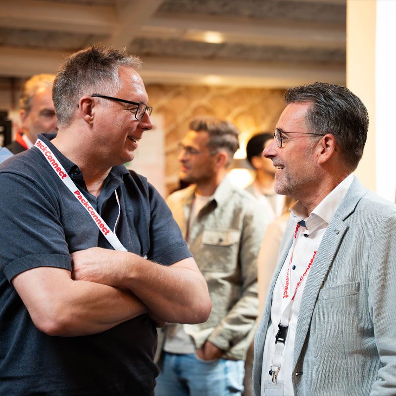 Two men engaged in conversation at a networking event, wearing name tag lanyards and surrounded by other attendees in a softly lit room.