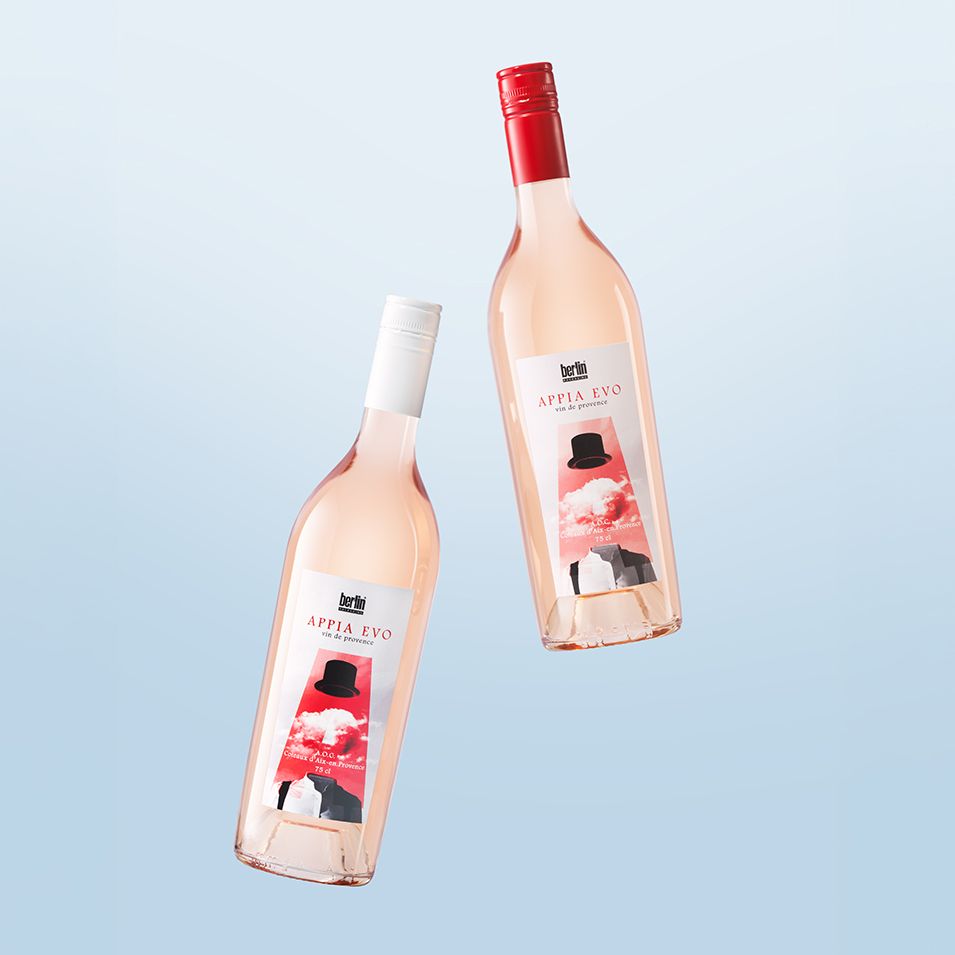 Two bottles of Appia Evo by Berlin Packaging filled with rosé wine with red and white caps, featuring a hat design on the label, set against a light blue background.