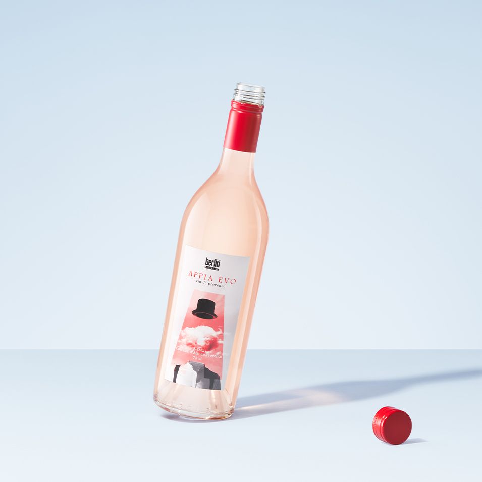 Appia Evo bottle by Berlin Packaging filled with rosé wine with a red screw cap lying beside it, set on a light blue surface against a light blue background.