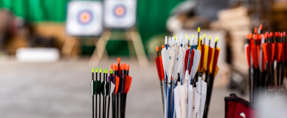 Pfeile in Behältern im Vordergrund, unscharfe Bogenschießscheiben im Hintergrund einer Indoor-Anlage