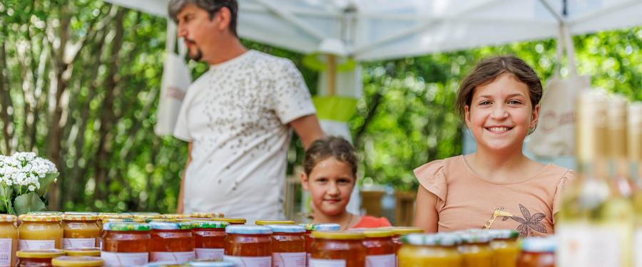 Lächelndes Mädchen und Kind stehen an einem Tisch mit verschiedenen Gläsern und Flaschen unter einem Pavillon auf einem Outdoor-Markt, Bäume im Hintergrund