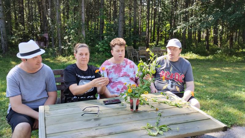 Vier Personen sitzen an einem Holztisch im Freien, halten Blumen in den Händen, umgeben von Wald und Natur.