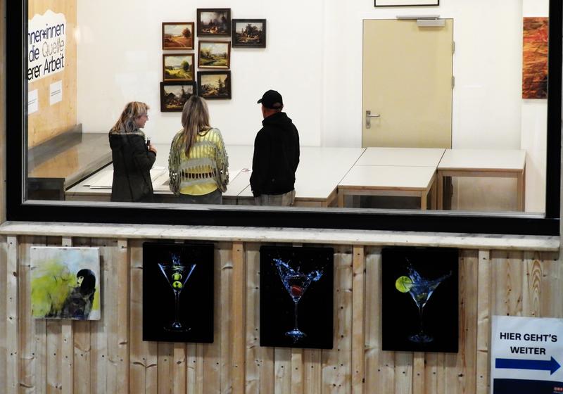 Drei Besucher betrachten Kunstwerke in einer Galerie; darunter hängen stilisierte Martini-Gemälde an einer Holzwand, daneben ein Hinweisschild mit Pfeil