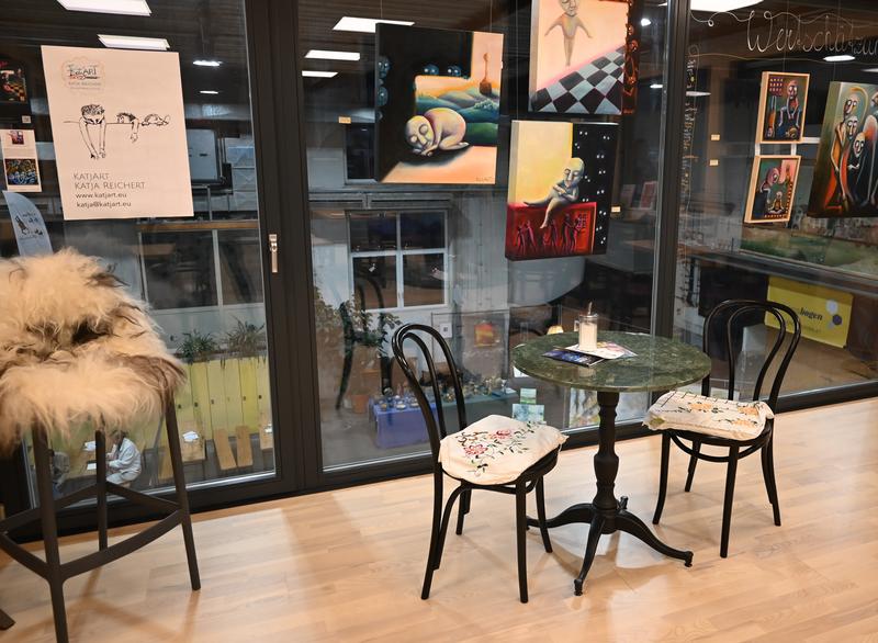 Gemütliche Café-Ecke mit zwei Stühlen und rundem Tisch, umgeben von farbenfroher Kunst und großen Fenstern; entspannte Atmosphäre
