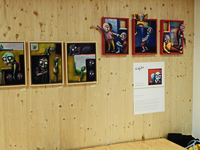 Kunstausstellung mit surrealen, farbenfrohen Gemälden auf einer Holzwand; Figuren mit großen Augen in unterschiedlichen Szenen