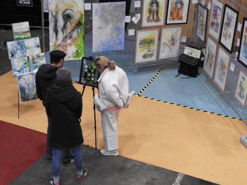 Zwei Personen betrachten Gemälde in einer Ausstellung; gerahmte Werke an Wänden und auf Staffeleien, Stativ in der Mitte
