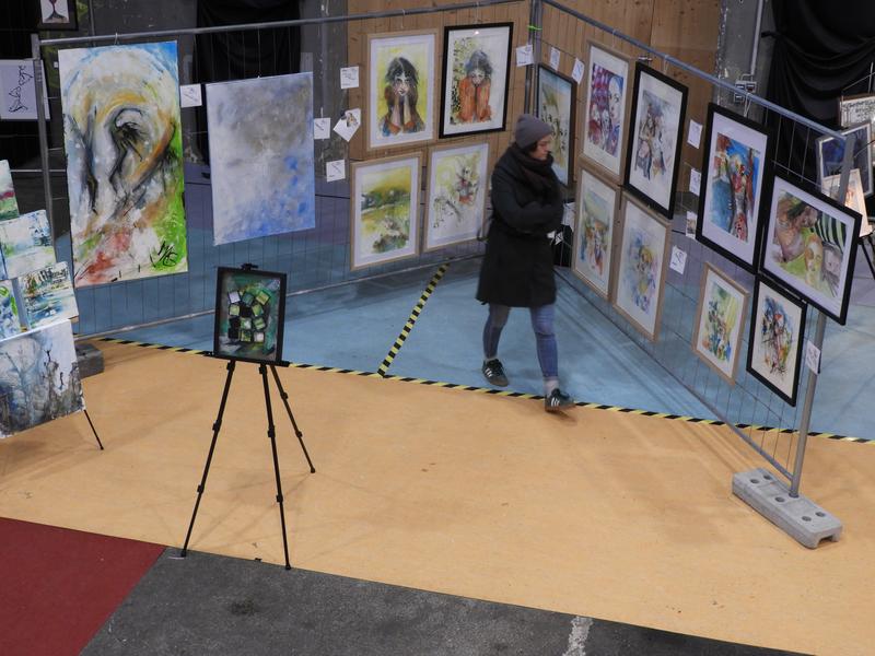 Person geht an einer Indoor-Ausstellung vorbei; Gemälde (Porträts und abstrakte Werke) auf Drahtgittern und Holzboden präsentiert