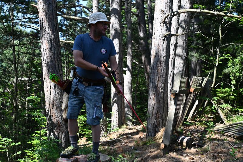 Mann im Wald mit Bogen und Pfeilen neben einer Zielscheibe; holt Pfeile im Sonnenlicht zwischen Bäumen