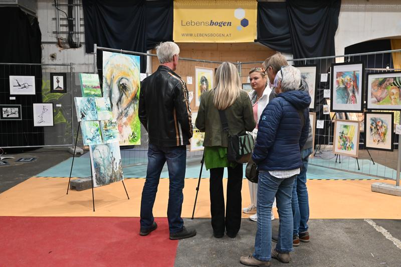 Besucher vor farbenfrohen Gemälden einer Ausstellung; Banner mit der Aufschrift „Lebensbogen“ im Hintergrund