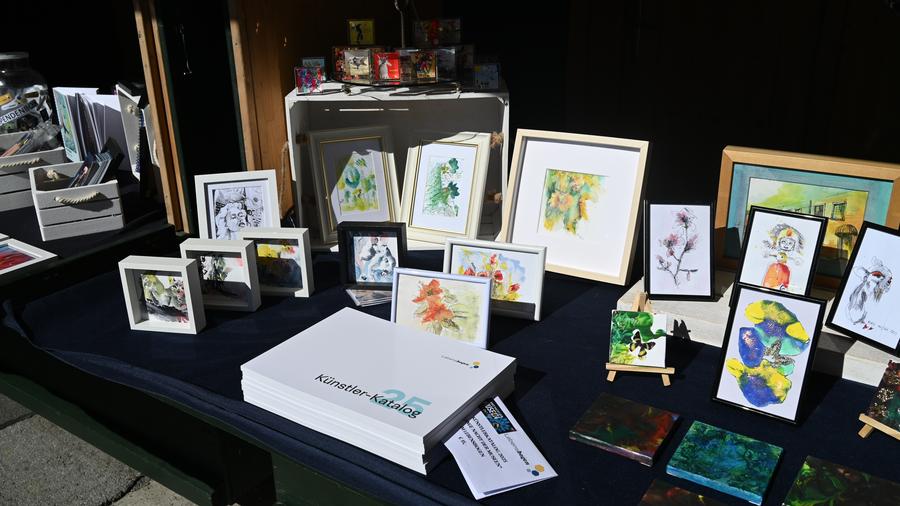 Gerahmte Kunstdrucke, farbenfrohe Gemälde und Künstlerutensilien auf einem schwarzen Tischtuch, präsentiert auf einem Outdoor-Markt