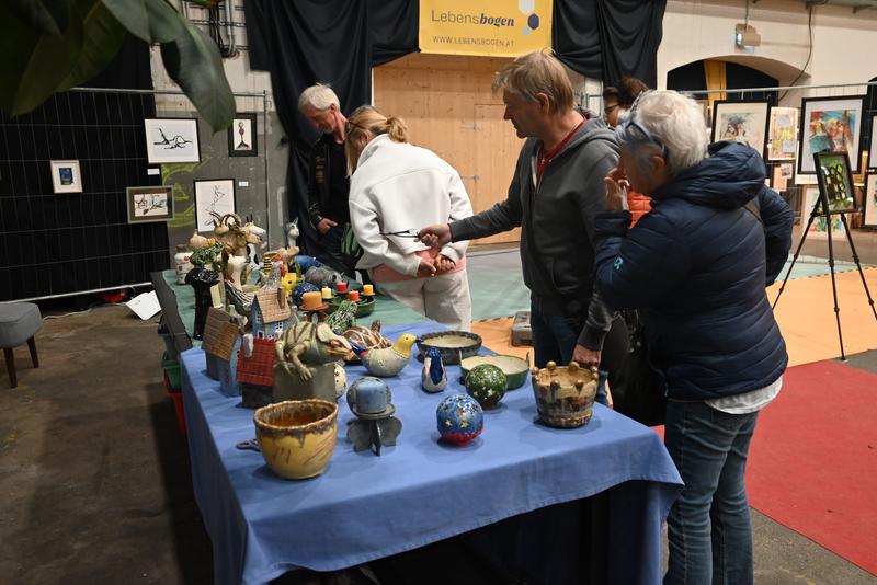 Besucher betrachten Keramikobjekte auf einem Tisch in einer Indoor-Ausstellung; gerahmte Kunstwerke im Hintergrund