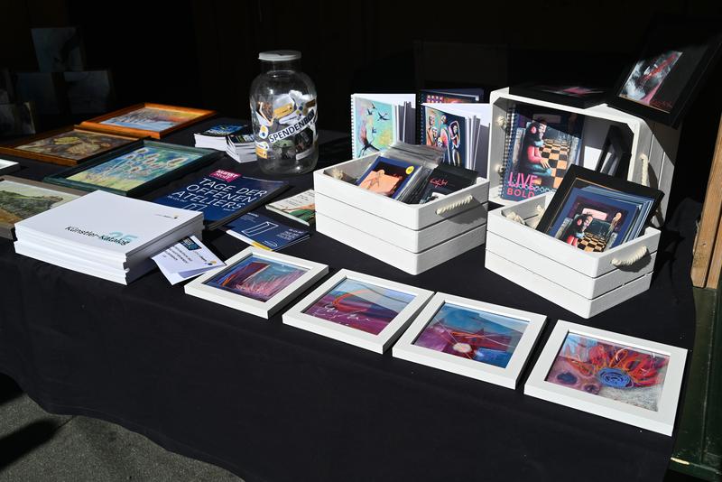 Präsentationstisch mit gerahmter Kunst, Spendenbox, Broschüren und verpackten Drucken auf schwarzem Tuch; Sonnenlicht betont die lebendigen Farben