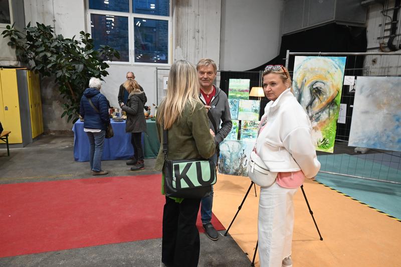 Besucher betrachten und besprechen Kunstwerke bei einer Indoor-Ausstellung; verschiedene Gemälde und Präsentationen im Hintergrund