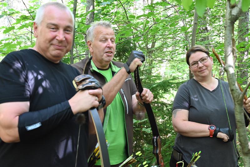 Drei Personen mit Bögen stehen lächelnd im Wald, in Freizeitkleidung mit Bogenschießausrüstung