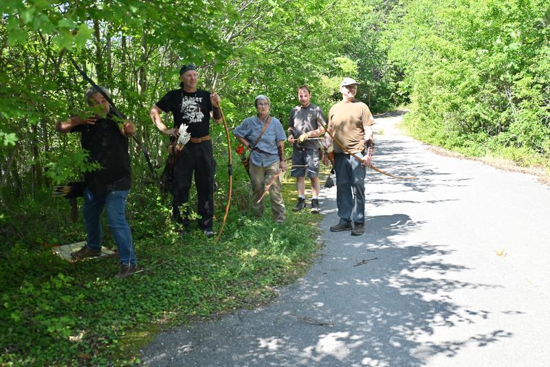 Fünf Personen mit Bögen stehen auf einem sonnigen Weg, umgeben von üppigem Grün; eine Person demonstriert das Bogenschießen