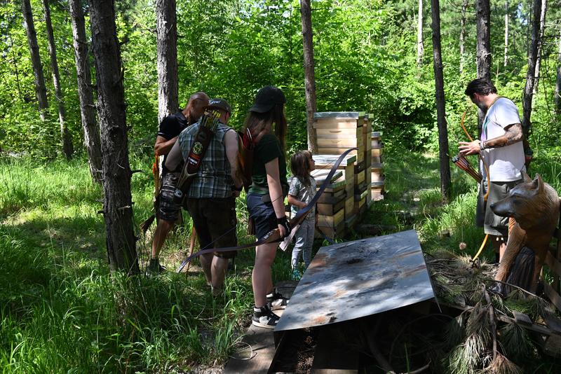 Gruppe mit Bögen im Wald bei einer Einführung ins Bogenschießen; Zielbereich und Tierfigur im Hintergrund