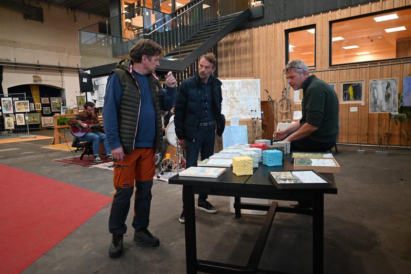 Drei Männer in einer Galerie; einer präsentiert Bücher auf einem Tisch, die anderen schauen zu; Kunstwerke im Hintergrund