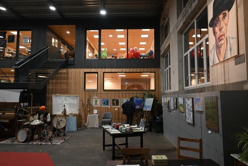 Galerieraum mit Gemälden an den Wänden, Schlagzeug und Büchern auf einem Tisch; eine Person steht nahe der Kunstwerke