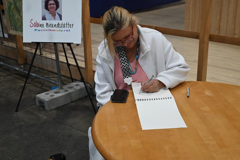 Frau sitzt an einem runden Holztisch und skizziert mit Bleistift; Poster mit ihrem Namen und Foto daneben