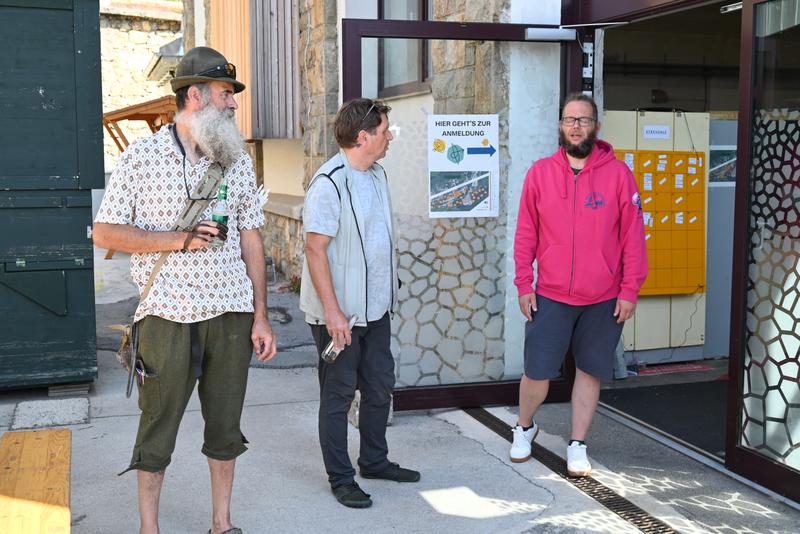 Drei Männer unterhalten sich vor einem Gebäude; einer bärtig in traditioneller Kleidung, ein anderer in Freizeitkleidung und der dritte im rosa Hoodie