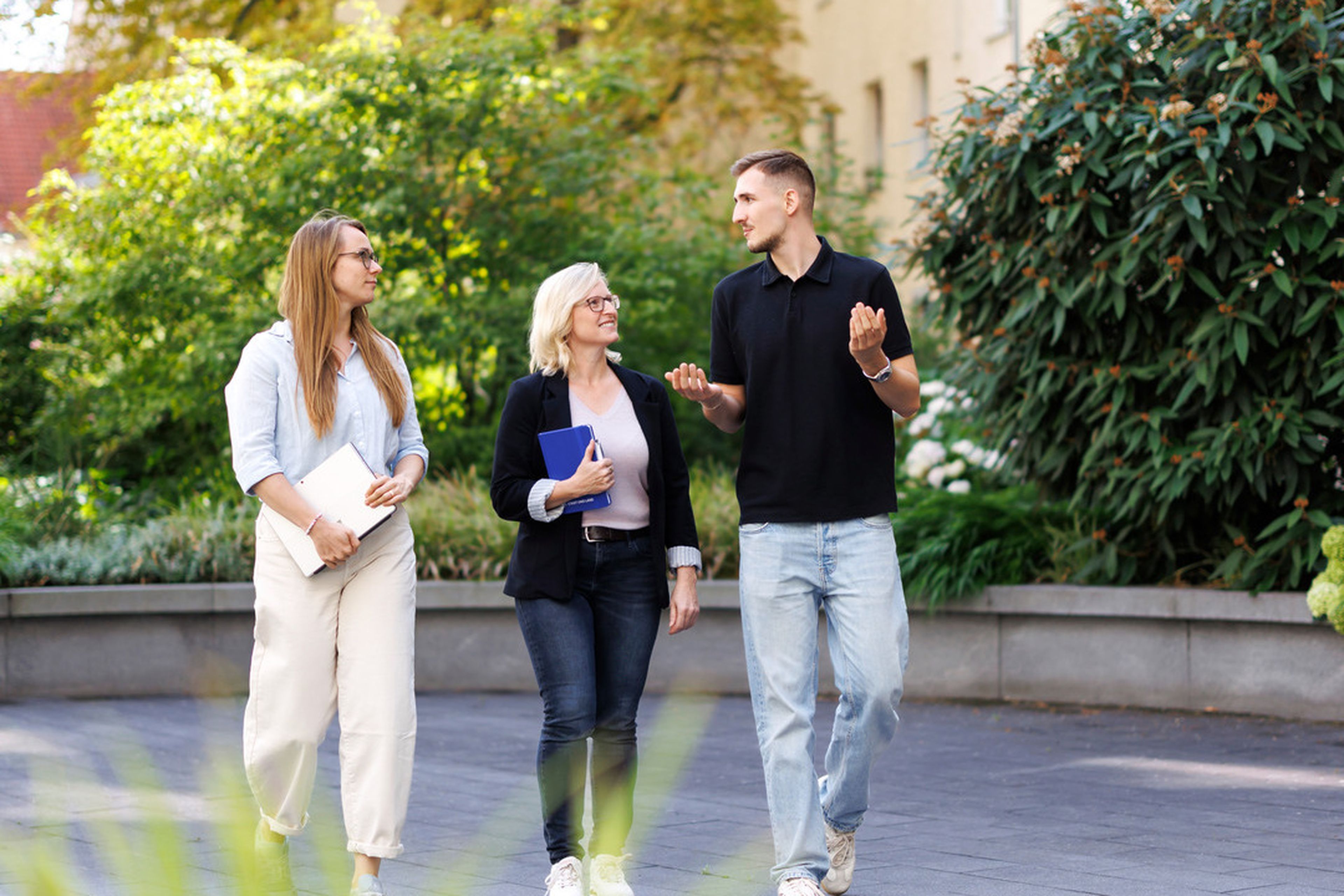Three people walking and talking outdoors near a building surrounded by greenery. They are holding notebooks and appear engaged in conversation.