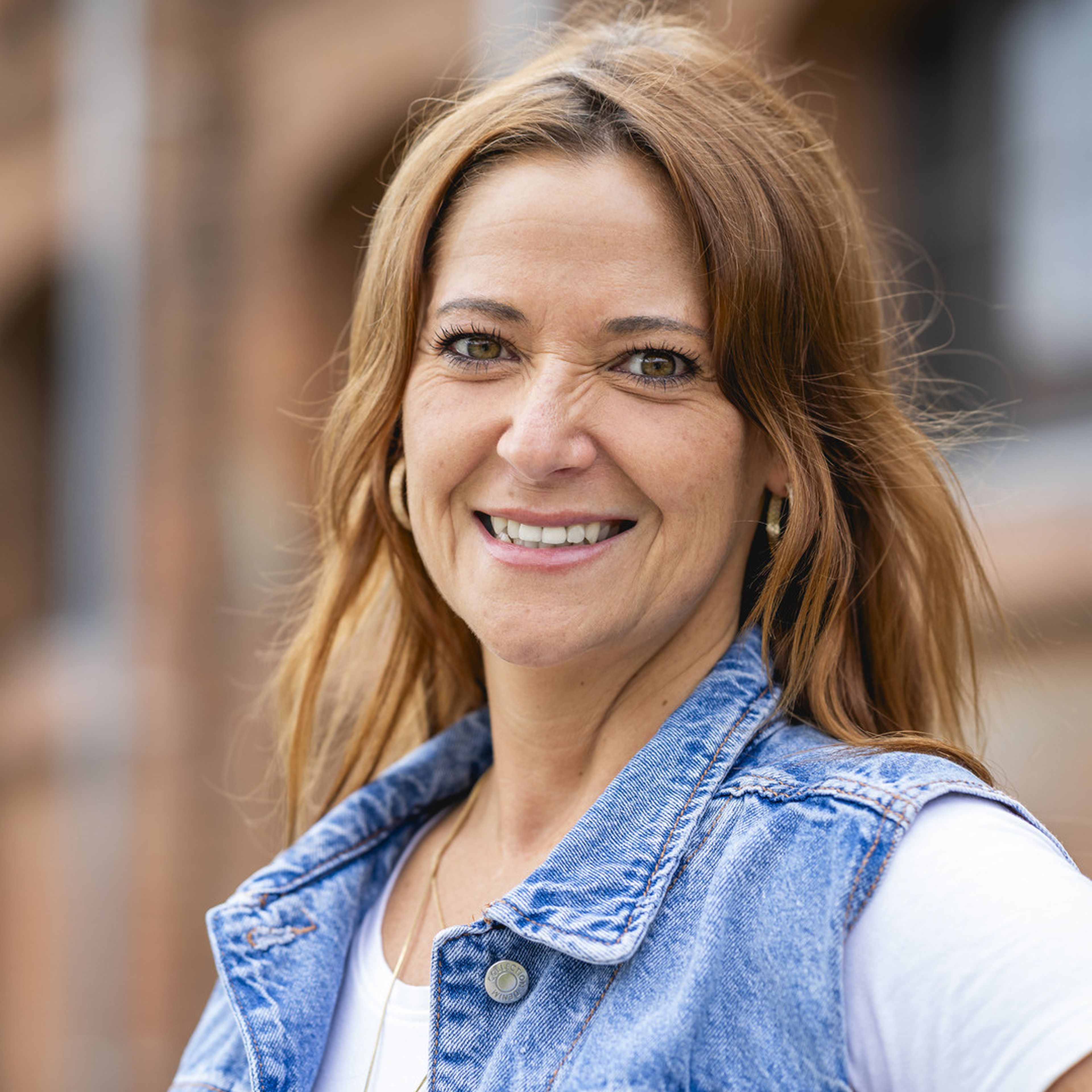 Smiling woman with long brown hair wearing a denim jacket and hoop earrings, standing outdoors with a blurred brick building in the background.