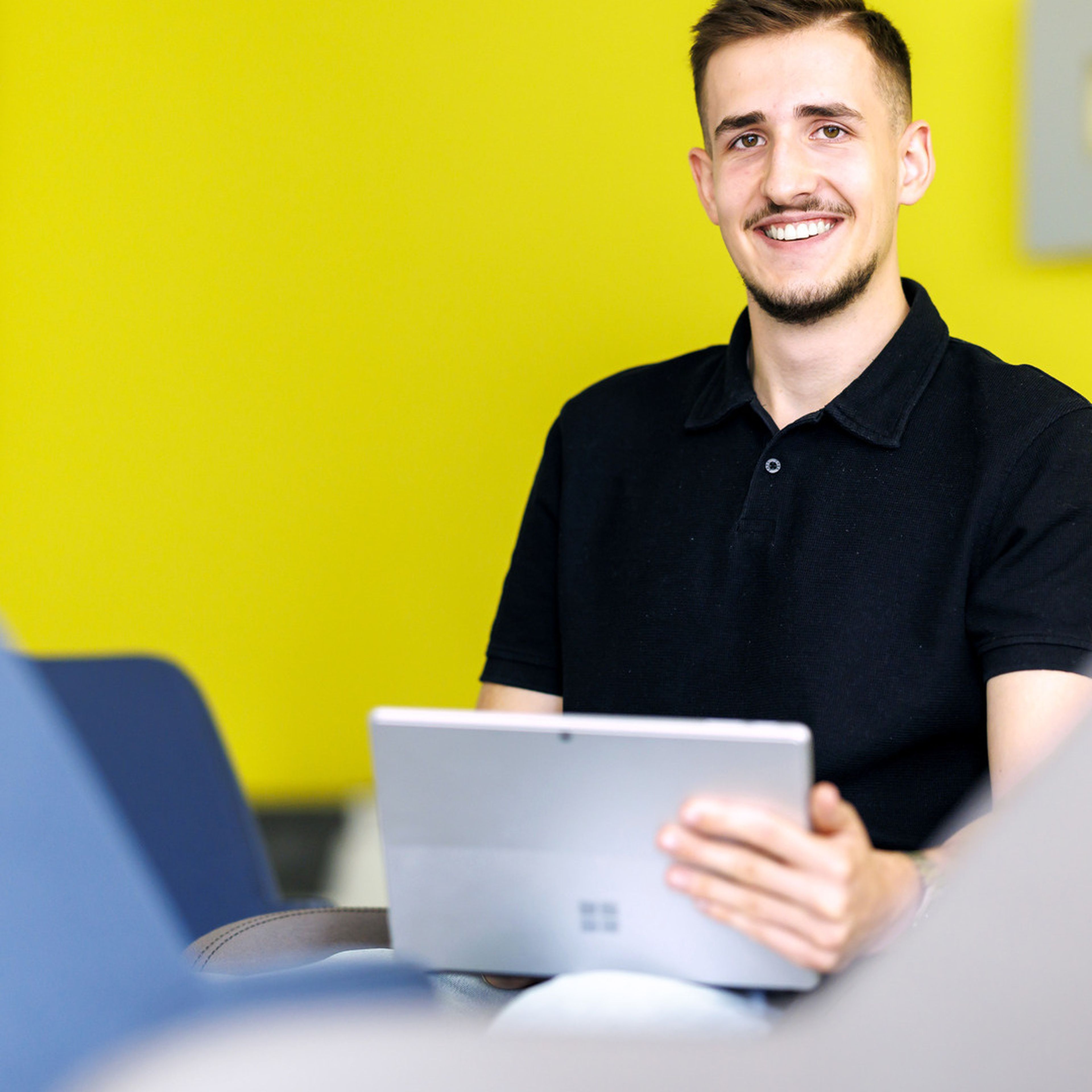 A smiling man in a black shirt holding a tablet, sitting against a bright yellow wall.