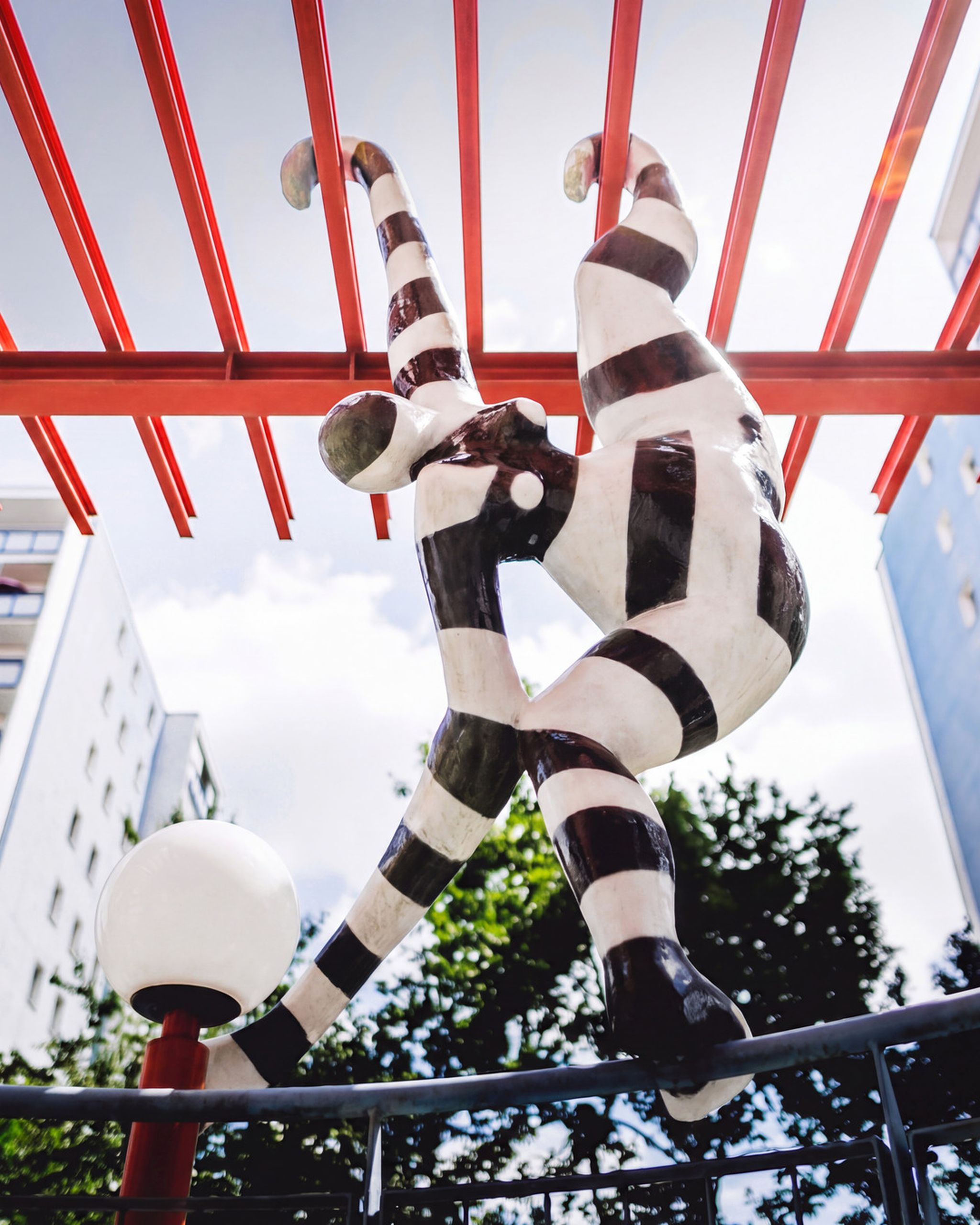 Sculpture of a whimsical figure with black and white stripes hanging upside down on a red structure, with buildings and trees in the background.