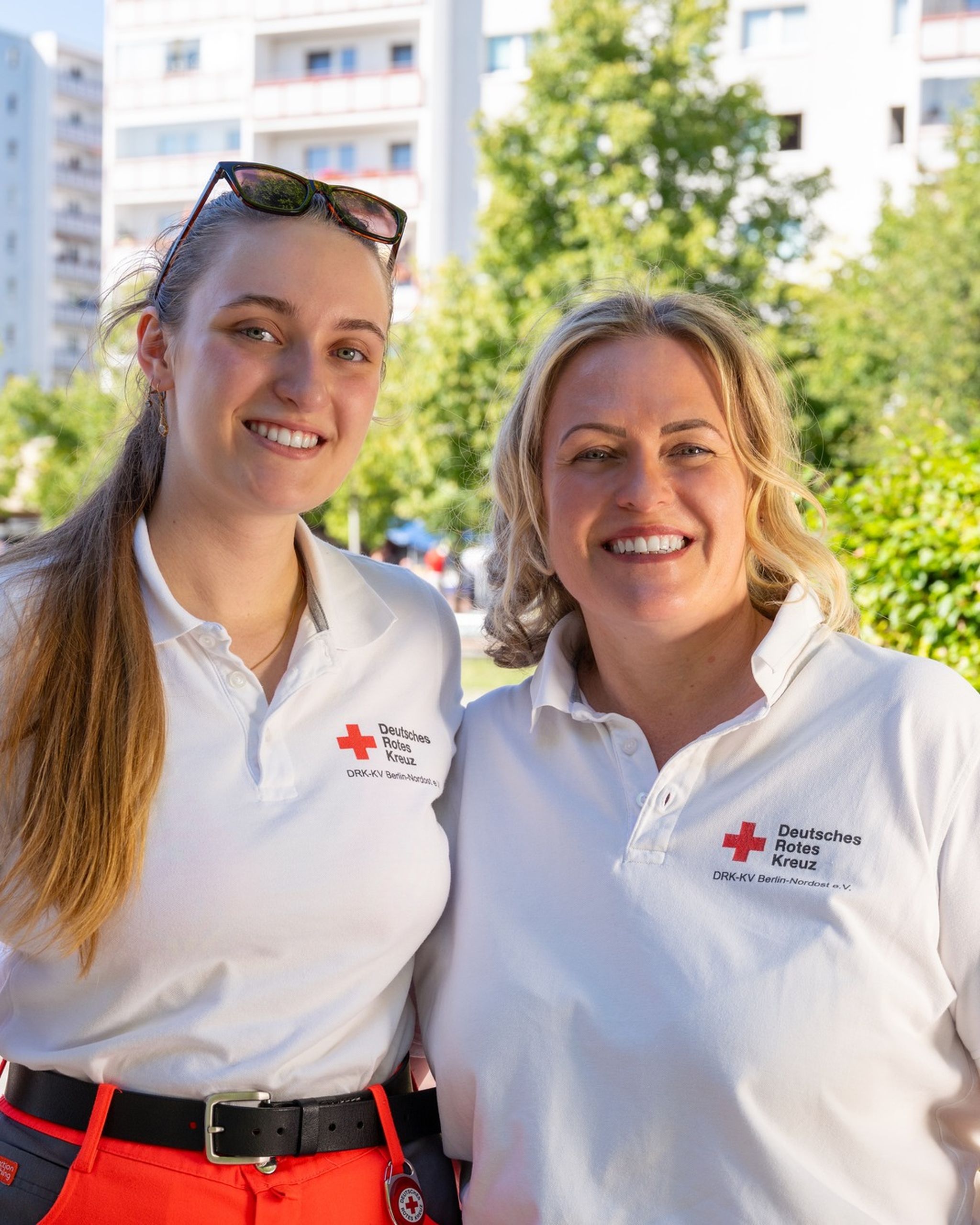 Two smiling women wearing matching Red Cross shirts, standing outdoors with buildings and trees in the background.