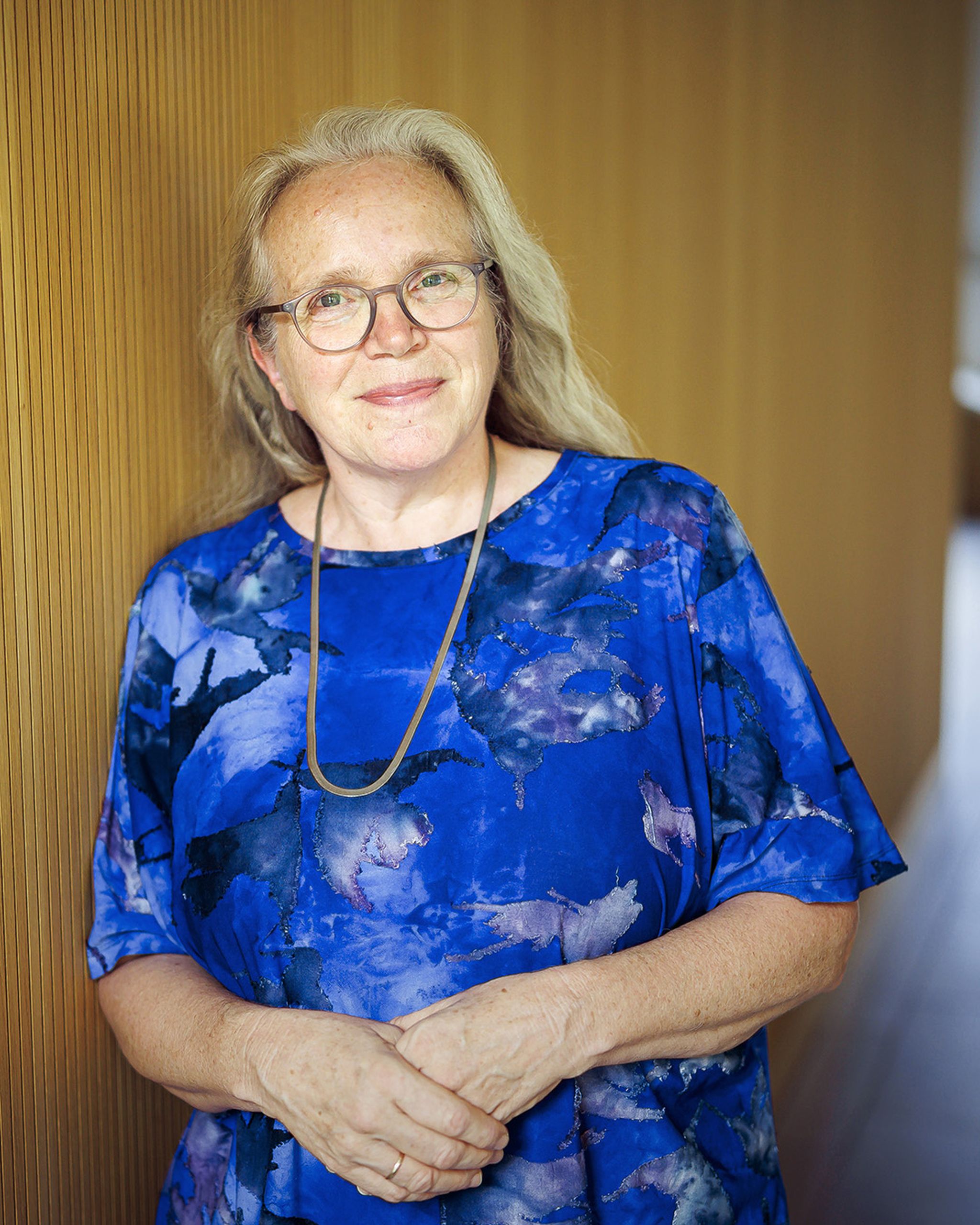 A woman with long hair and glasses, wearing a blue patterned blouse, stands against a wooden wall, smiling with hands clasped.