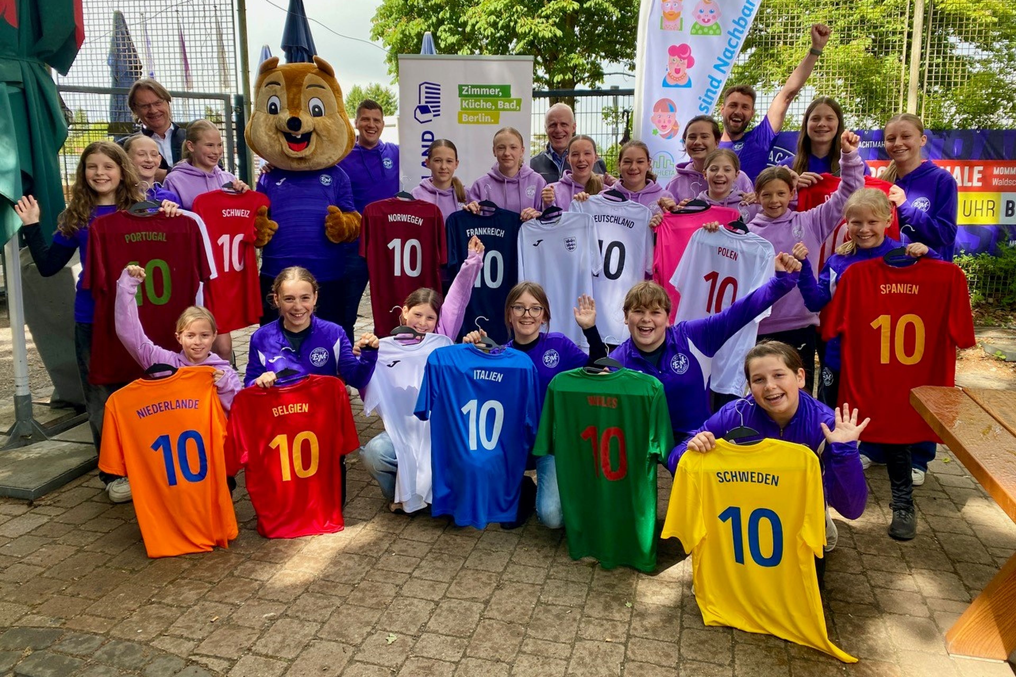 Das Mädchenteam hält Fußballtrikots mit Ländernamen und der Nummer 10, neben einem Maskottchen im Freien.
