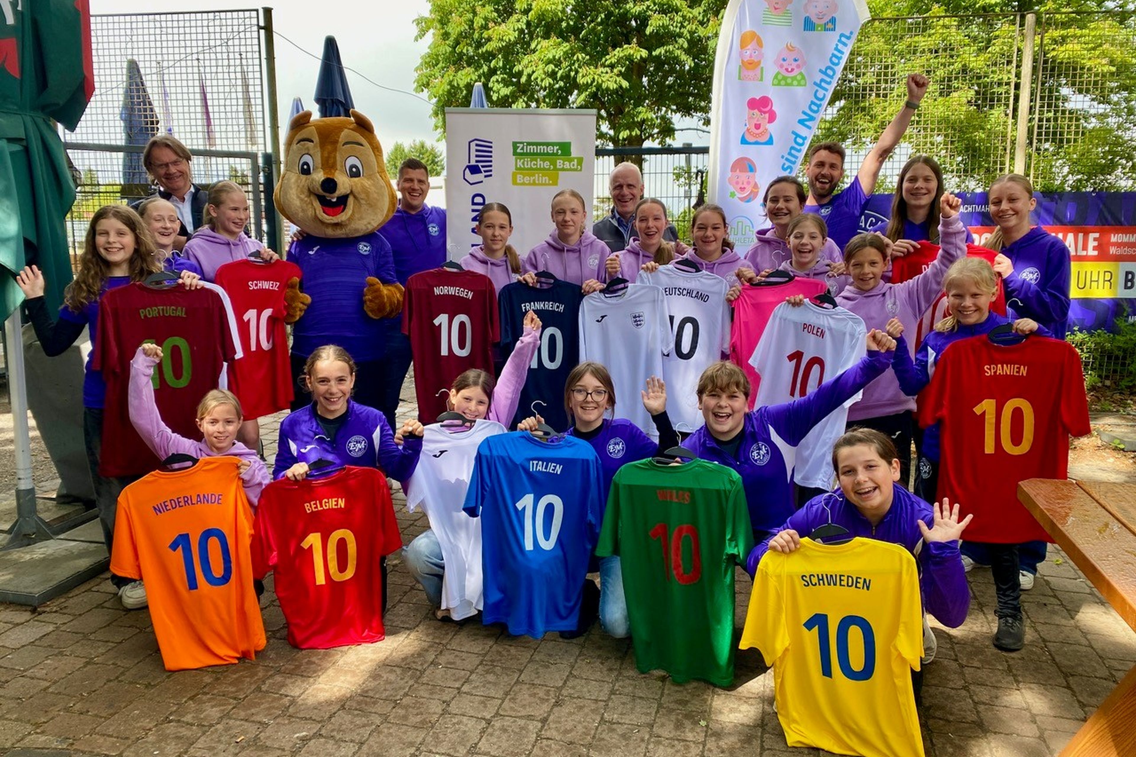Eine Gruppe von Kindern in lila Hoodies hält bunte Fußballtrikots mit Länderflaggen, neben einem Maskottchen.