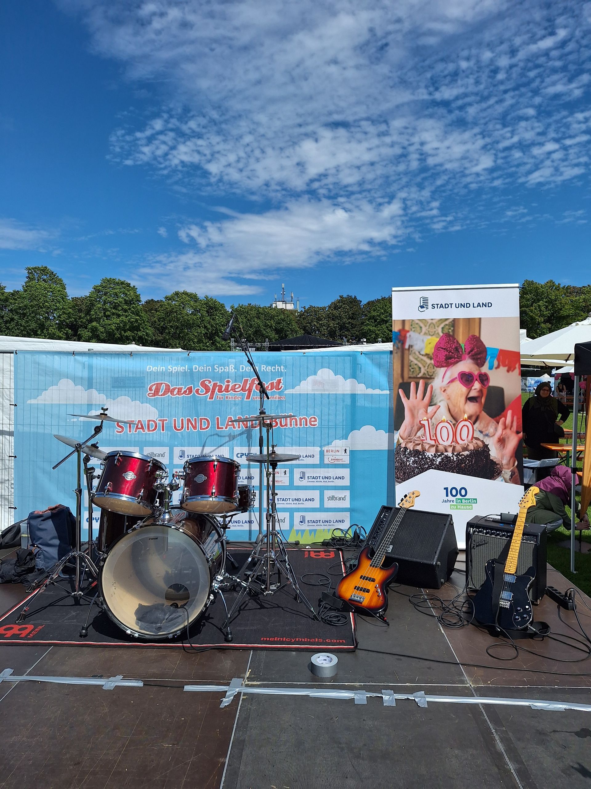 Ein Schlagzeug und zwei Gitarren auf einer Bühne im Freien, mit einem Werbebanner im Hintergrund.