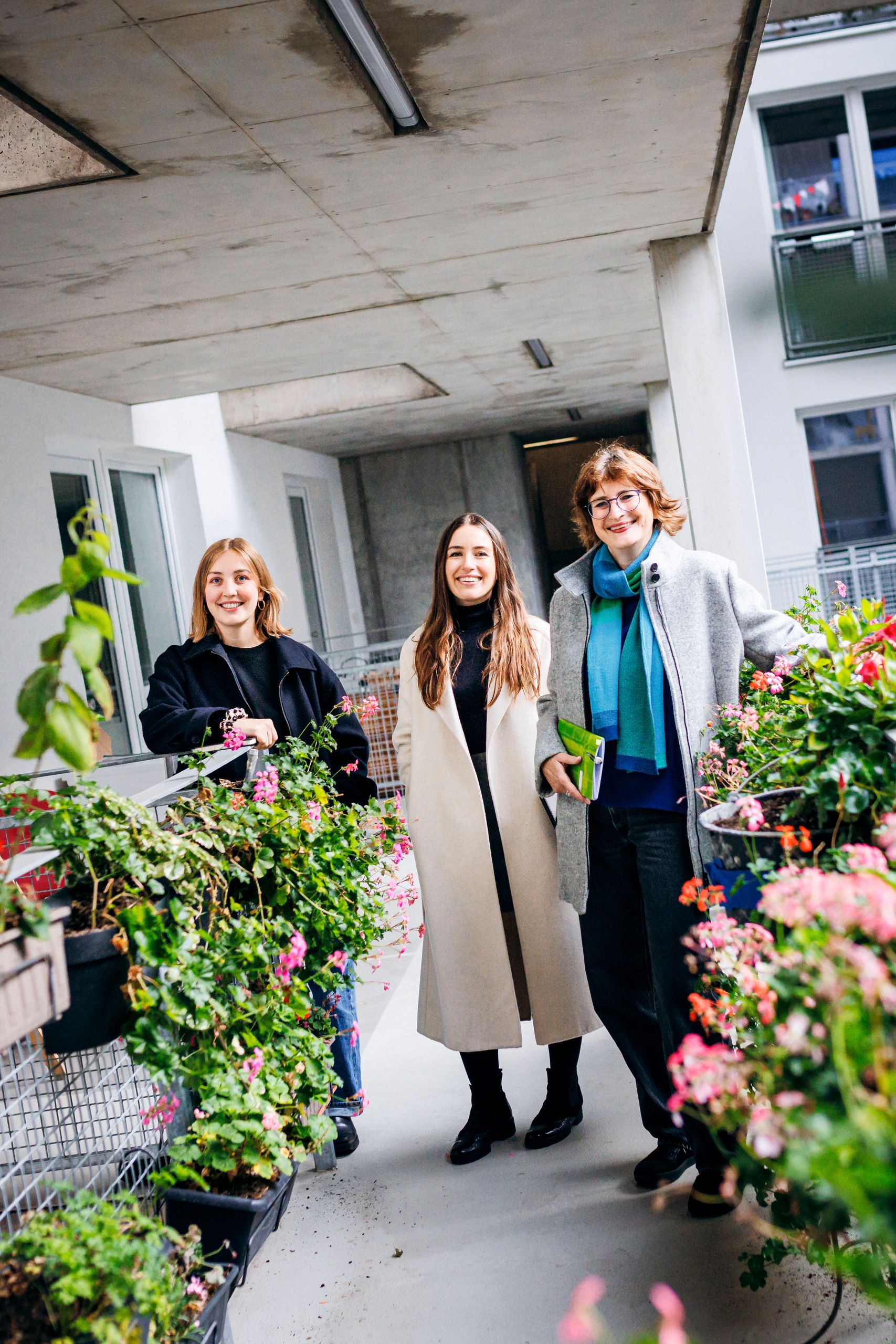 Drei Frauen stehen lächelnd auf einem Balkon mit blühenden Pflanzen, umgeben von modernen Gebäuden.
