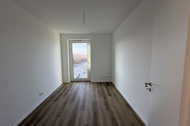 Leeres Zimmer mit Holzfußboden, weiß gestrichenen Wänden und einer Glastür, die zu einem Balkon führt.