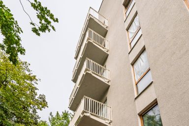 A beige building facade with several balconies, viewed from below, next to leafy green trees against a clear sky.