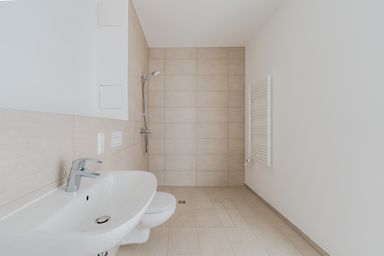 Minimalist bathroom with beige tiled floor and walls, featuring a white sink, shower, toilet, and wall-mounted towel heater.