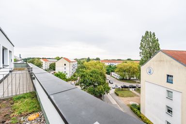 Balcony view of residential neighborhood with red-roofed buildings, trees, and parked cars on a cloudy day.