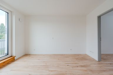 Empty room with light wooden floors, white walls, a large window on the left, and an open doorway on the right.