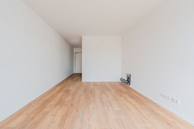 Leerer Raum mit hellem Holzfußboden und weißen Wänden, Blick auf eine offene Tür im Hintergrund.