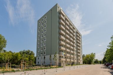 Elfgeschossiges Wohngebäude mit Balkonen, umgeben von Bäumen und einem Parkplatz, unter blauem Himmel.