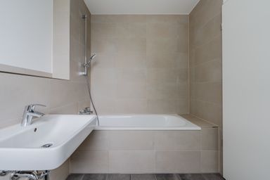 Modern bathroom with beige tiles, featuring a white bathtub and a mounted hand shower. A sleek, rectangular sink is visible nearby.