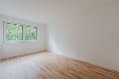 Empty room with light wood flooring, white walls, and large windows showing green trees outside.
