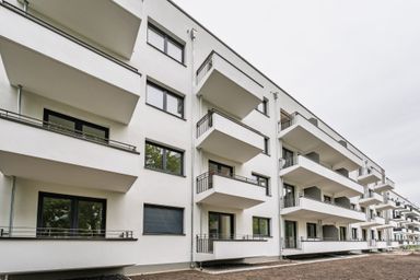Modern apartment building with white facade and multiple balconies, featuring large windows and a simple, contemporary design under a cloudy sky.