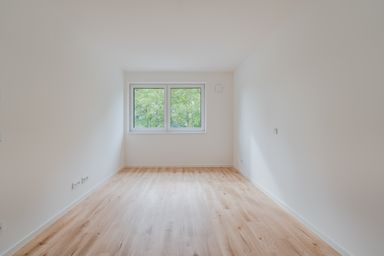 Empty room with light wood flooring and white walls, featuring a large window with a view of green trees outside.
